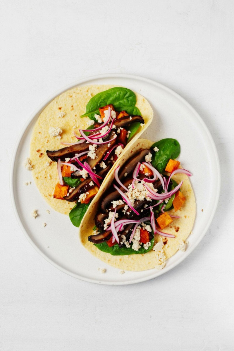 An overhead image of two neatly folded vegan portobello mushroom tacos, which are garnished with thinly sliced pickled red onion and plant-based "feta" cheese.